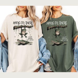 May include: Two t-shirts, one cream and one olive green, featuring a whimsical illustration. The design shows a frog hanging from a branch above an alligator, with the text "HANG IN THERE" and "could always get worse".