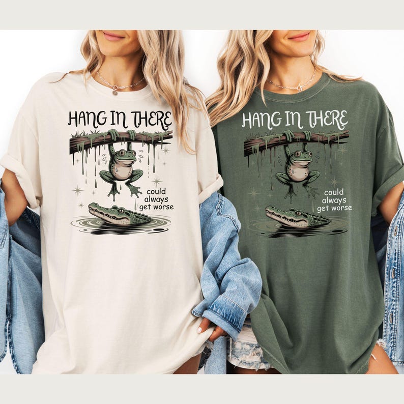 May include: Two t-shirts, one cream and one olive green, featuring a whimsical illustration. The design shows a frog hanging from a branch above an alligator, with the text "HANG IN THERE" and "could always get worse".
