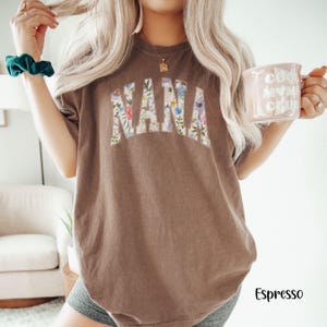May include: Espresso-colored t-shirt with the word "NANA" in a floral design across the front. The shirt is a relaxed fit and has short sleeves. A mug with the text "cool mom club" is held in the right hand.