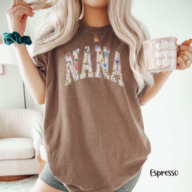 May include: Espresso-colored t-shirt with the word "NANA" in a floral design across the front. The shirt is a relaxed fit and has short sleeves. A mug with the text "cool mom club" is held in the right hand.