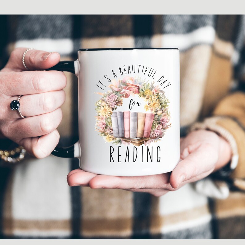 May include: White ceramic mug with a black handle and rim, featuring the text "IT'S A BEAUTIFUL DAY for READING". The design includes a floral wreath surrounding a stack of books in pastel colors. A perfect gift for book lovers.
