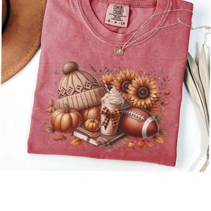 May include: A red t-shirt with a fall-themed design featuring a pumpkin, a coffee cup, a football, a knitted hat, and sunflowers. The text "COMFORT COLORS" is visible on the tag.
