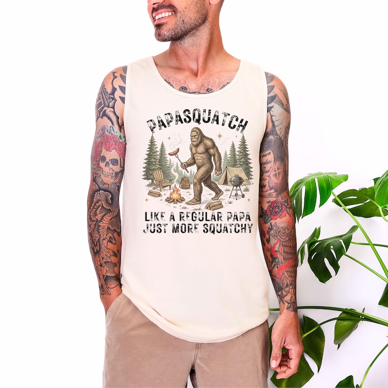 May include: Cream vest top featuring a Bigfoot graphic with a sausage over a campfire. Text reads "Papasquatch Like a Regular Papa Just More Squatchy."