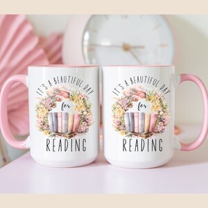 May include: Two white ceramic mugs with pink handles and interiors. Each mug features a floral wreath design with the text "It's a beautiful day for READING". The design includes a stack of books.