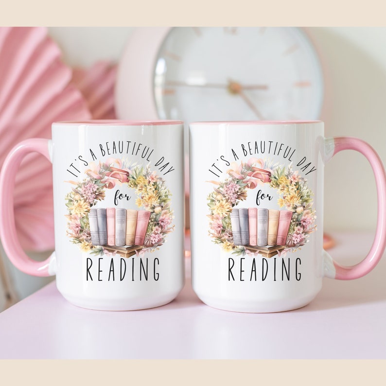 May include: Two white ceramic mugs with pink handles and interiors. Each mug features a floral wreath design with the text "It's a beautiful day for READING". The design includes a stack of books.