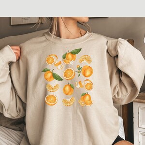May include: A beige sweatshirt with a graphic print of oranges and orange slices. The oranges are illustrated in a watercolor style with green leaves and white flowers.