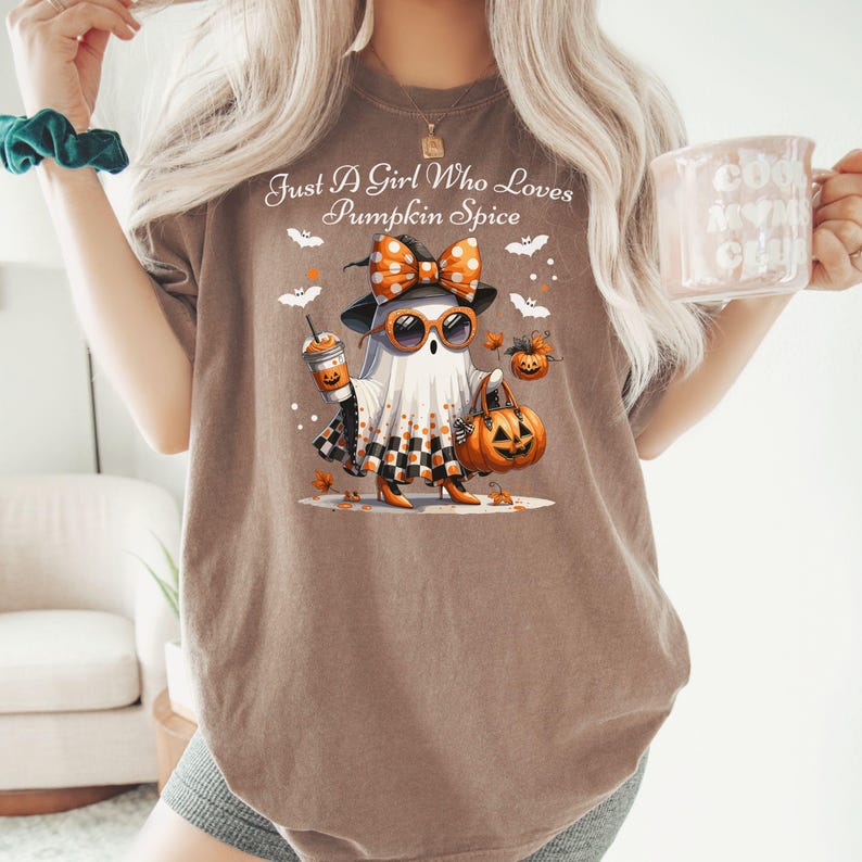 May include: Brown t-shirt with the text "Just A Girl Who Loves Pumpkin Spice." The shirt features a cartoon ghost wearing sunglasses, a bow, and holding a pumpkin spice latte and a jack-o'-lantern bag. Bats and pumpkins are also in the design.
