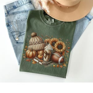 May include: A green t-shirt with a graphic print of a brown and white knit hat, pumpkins, sunflowers, a football, and a coffee drink with whipped cream. The graphic is surrounded by fall leaves.
