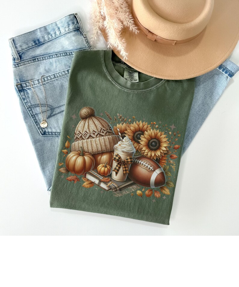 May include: A green t-shirt with a graphic print of a brown and white knit hat, pumpkins, sunflowers, a football, and a coffee drink with whipped cream. The graphic is surrounded by fall leaves.
