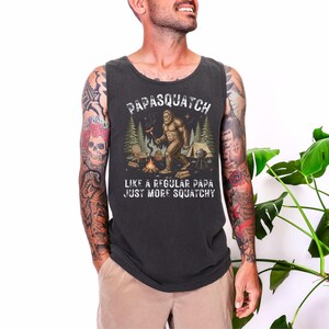 May include: Dark grey vest top featuring the text "Papasquatch" above a Bigfoot illustration near a campfire. The text below reads "Like a regular papa just more squatchy."