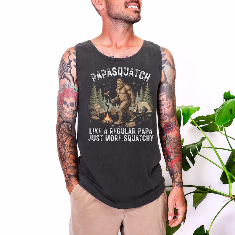 May include: Dark grey vest top featuring the text "Papasquatch" above a Bigfoot illustration near a campfire. The text below reads "Like a regular papa just more squatchy."