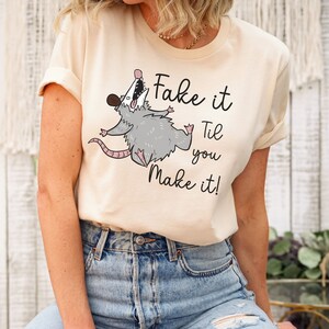 Puede incluir: Una camiseta beige con un opossum de dibujos animados y el texto "Fake it Til you Make it!"