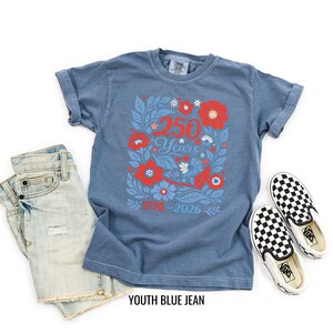 May include: A blue t-shirt with a floral design featuring the text "250 Years 1776-2026". The shirt is paired with denim shorts and black and white checkered slip-on shoes. The words "YOUTH BLUE JEAN" are printed at the bottom.