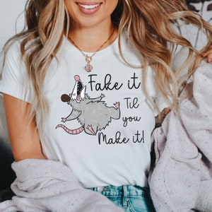 Puede incluir: Camiseta blanca con un opossum de dibujos animados y el texto "Fake it Til you Make it!"
