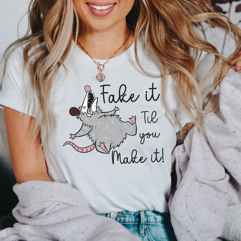 Puede incluir: Camiseta blanca con un opossum de dibujos animados y el texto "Fake it Til you Make it!"