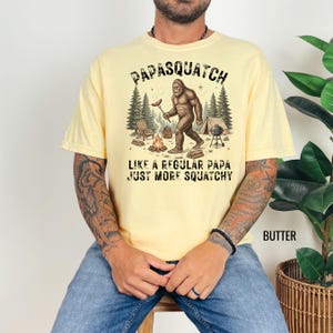 May include: Pale yellow t-shirt featuring a Bigfoot illustration with a sausage near a campfire. The text above reads "PAPASQUATCH" and below "LIKE A REGULAR PAPA JUST MORE SQUATCHY".