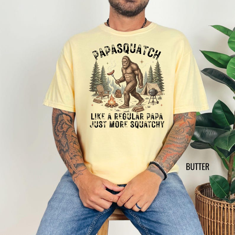 May include: Pale yellow t-shirt featuring a Bigfoot illustration with a sausage near a campfire. The text above reads "PAPASQUATCH" and below "LIKE A REGULAR PAPA JUST MORE SQUATCHY".