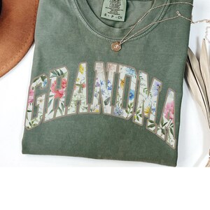 May include: Green t-shirt with the word "Grandma" in large, white letters outlined in brown. The letters are filled with a floral pattern.