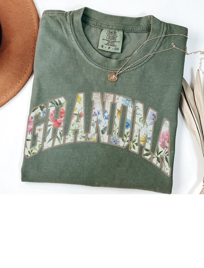 May include: Green t-shirt with the word "Grandma" in large, white letters outlined in brown. The letters are filled with a floral pattern.