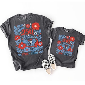 May include: Two charcoal gray t-shirts with a floral design in red, white, and blue. The design includes the text "250 Years" and the dates "1776-2026". A pair of black and white checkered slip-on shoes are in the foreground.