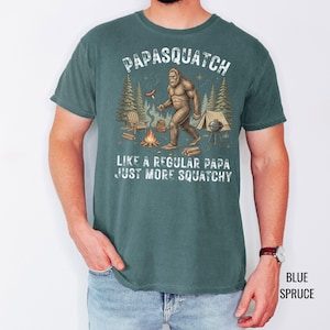 May include: Blue spruce t-shirt featuring a Bigfoot illustration cooking over a campfire. The text reads "Papasquatch, Like a regular papa, just more squatchy."
