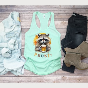May include: A light blue tank top with a watercolor illustration of a raccoon holding a beer mug. The raccoon is surrounded by a yellow and orange sunburst. The text "PROST!" is printed in colorful letters below the raccoon.