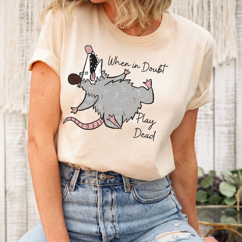 Pu&ograve; includere: Una maglietta beige con un disegno di opossum dei cartoni animati e il testo "When in doubt, play dead".