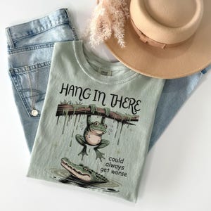 May include: A light green t-shirt with the phrase "Hang in There" and a cartoon frog hanging from a branch above an alligator. The shirt is paired with blue jeans and a tan hat.