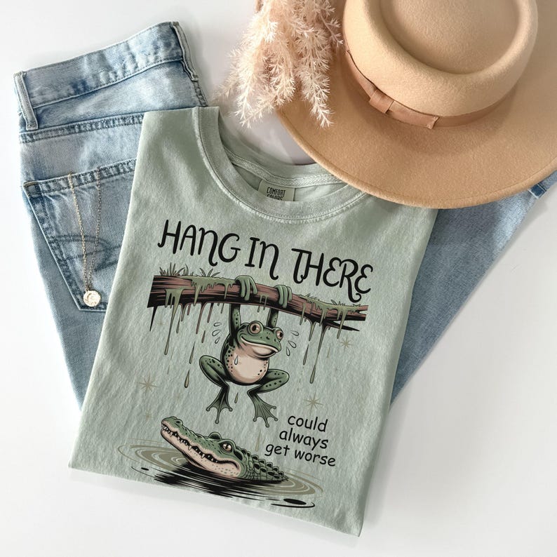 May include: A light green t-shirt with the phrase "Hang in There" and a cartoon frog hanging from a branch above an alligator. The shirt is paired with blue jeans and a tan hat.