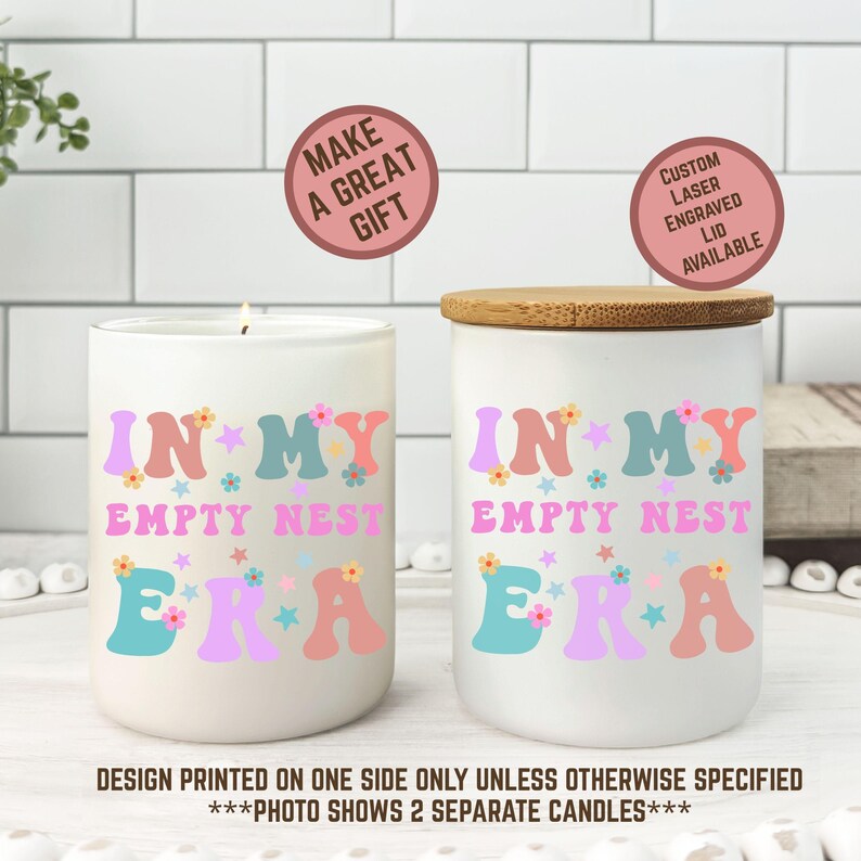 Op de afbeelding: Twee witte keramische kaarsen met de tekst "IN MY EMPTY NEST ERA" in kleurrijke letters in retrostijl. E&eacute;n kaars heeft een aangestoken lont, de andere heeft een houten deksel. De afbeelding bevat de tekst "MAKE A GREAT GIFT" en "CUSTOM LASER ENGRAVED LID AVAILABLE".