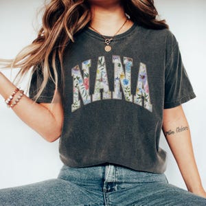 May include: Dark gray t-shirt with the word "NANA" in a floral design. The letters are filled with a variety of colorful flowers and green leaves. The shirt has a vintage, worn look.