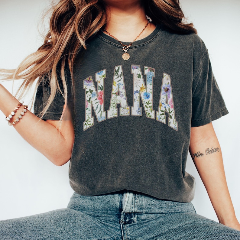 May include: Dark gray t-shirt with the word "NANA" in a floral design. The letters are filled with a variety of colorful flowers and green leaves. The shirt has a vintage, worn look.