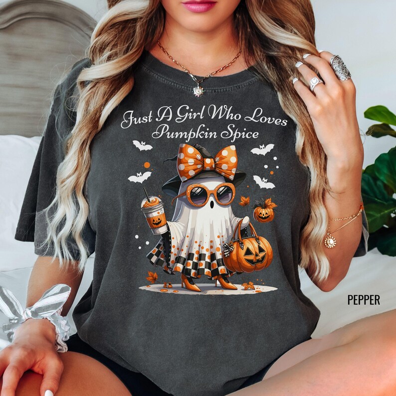 May include: Dark gray t-shirt with the text "Just A Girl Who Loves Pumpkin Spice." The graphic features a ghost wearing sunglasses, a bow, and holding a pumpkin spice latte and a jack-o'-lantern bag. Bats and pumpkins are also in the design.