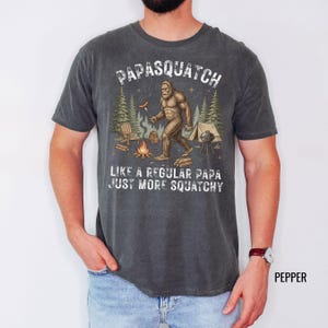 May include: Dark grey t-shirt featuring a Bigfoot graphic with the text "PAPASQUATCH LIKE A REGULAR PAPA JUST MORE SQUATCHY". The design shows a creature grilling a sausage. The shirt has a faded, vintage appearance.