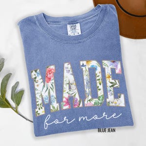 May include: A blue jean Comfort Colors t-shirt with the word "MADE" in large floral print letters. Below the word "MADE" is the phrase "for more" in cursive. The shirt is a crew neck and has a tag that says "Comfort Colors".