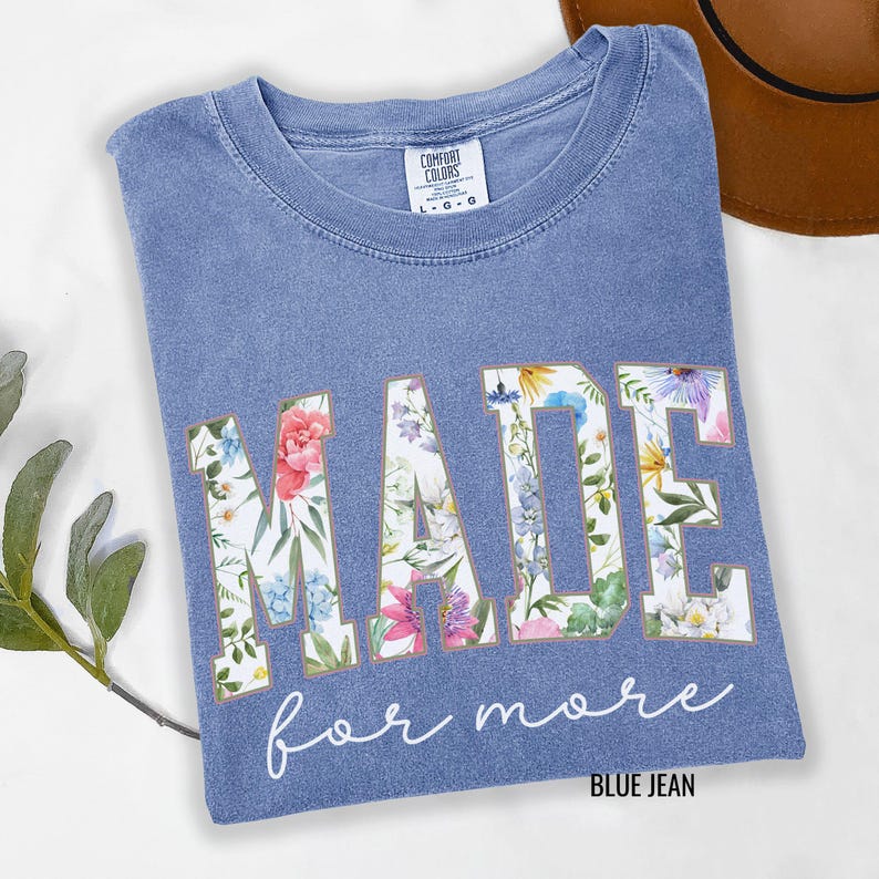 May include: A blue jean Comfort Colors t-shirt with the word "MADE" in large floral print letters. Below the word "MADE" is the phrase "for more" in cursive. The shirt is a crew neck and has a tag that says "Comfort Colors".