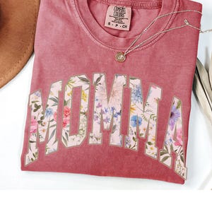 May include: A red t-shirt with a floral print design that says "MOMMA" in a large, white, outlined font.