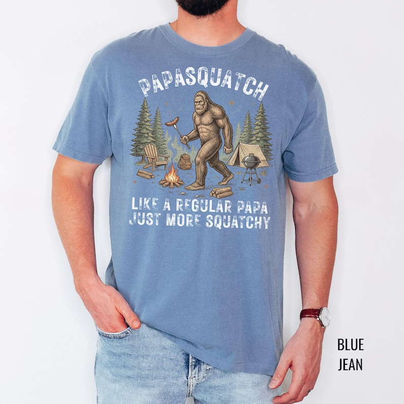 May include: A blue t-shirt featuring the words "Papasquatch" and a Bigfoot graphic holding a hot dog. The design includes a campfire, tent, and trees. The text below reads "Like a regular papa, just more squatchy."