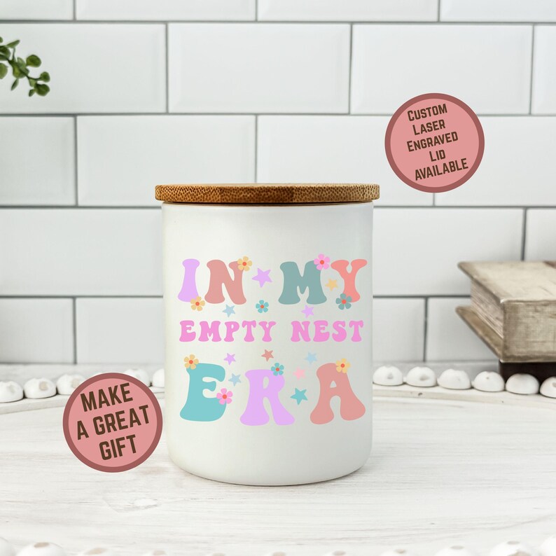 Op de afbeelding: Een witte keramische pot met een houten deksel, met de tekst "IN MY EMPTY NEST ERA" in kleurrijke letters in retrostijl. De pot heeft een ronde sticker met de tekst "MAKE A GREAT GIFT". Een tweede sticker zegt "CUSTOM LASER ENGRAVED LID AVAILABLE".