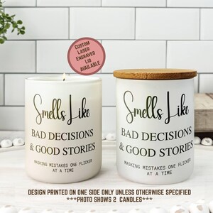 May include: Two white candles with the text "Smells Like Bad Decisions & Good Stories" in black script. One candle has a lit wick, the other has a wooden lid. The text "Masking mistakes one flicker at a time" is also printed on the candles.