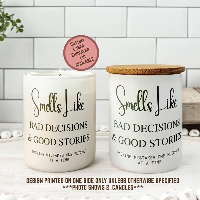 May include: Two white candles with the text "Smells Like Bad Decisions & Good Stories" in black script. One candle has a lit wick, the other has a wooden lid. The text "Masking mistakes one flicker at a time" is also printed on the candles.
