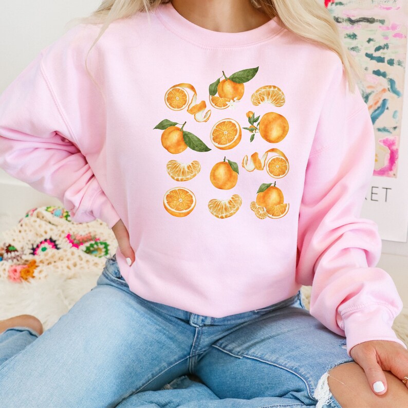 May include: A pink sweatshirt with a graphic print of oranges and orange slices. The oranges are illustrated in a watercolor style.