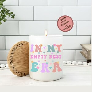 Op de afbeelding: Een witte kaars met de tekst "IN MY EMPTY NEST ERA" in pastelkleuren. De kaars heeft een houten deksel met gegraveerde tekst. Een sticker leest "Custom Laser Engraved Lid Available".