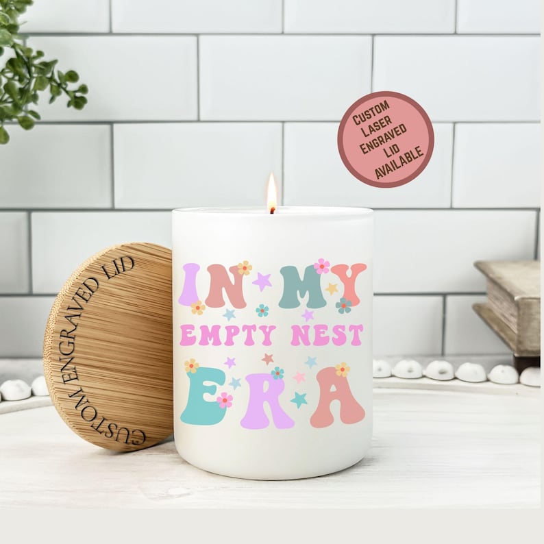 Op de afbeelding: Een witte kaars met de tekst "IN MY EMPTY NEST ERA" in pastelkleuren. De kaars heeft een houten deksel met gegraveerde tekst. Een sticker leest "Custom Laser Engraved Lid Available".