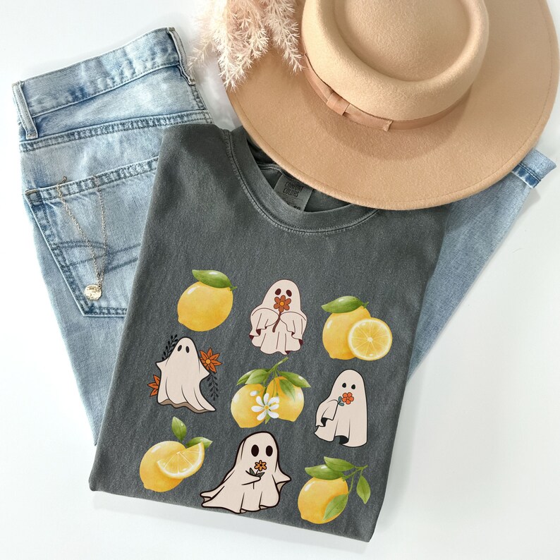 May include: A dark gray t-shirt with a graphic print of ghosts holding lemons. The ghosts are white and the lemons are yellow. The shirt is folded and laid on a pair of blue jeans and a brown hat.