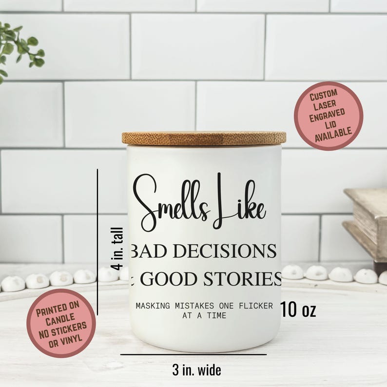 May include: A white candle with a bamboo lid, measuring 4 inches tall and 3 inches wide. The candle has the text "Smells Like Bad Decisions: Good Stories" printed on it. The candle is 10 oz and has the text "Masking mistakes one flicker at a time".