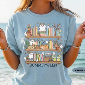 May include: Light blue t-shirt with a whimsical illustration of bookshelves filled with books, ghosts, and animals. The word "SUMMERWEEN" is printed below the shelves. The shirt is a chambray color.