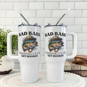 May include: Two white insulated tumblers with handles and lids. Each tumbler features a graphic of a bass wearing a hat and sunglasses, with the text "BAD BASS FISHING CLUB GET HOOKED". Stainless steel straws are inserted into the lids.