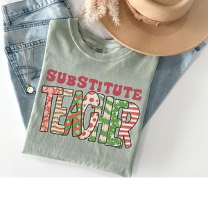 May include: A light green t-shirt with the text "Substitute Teacher" in red and green letters. The letters are decorated with a Christmas-themed pattern.