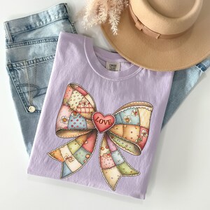 May include: A lilac-colored t-shirt featuring a colorful patchwork bow design with a red heart that says "LOVE". The shirt is laid on top of a pair of light blue jeans, with a tan hat and a gold necklace.